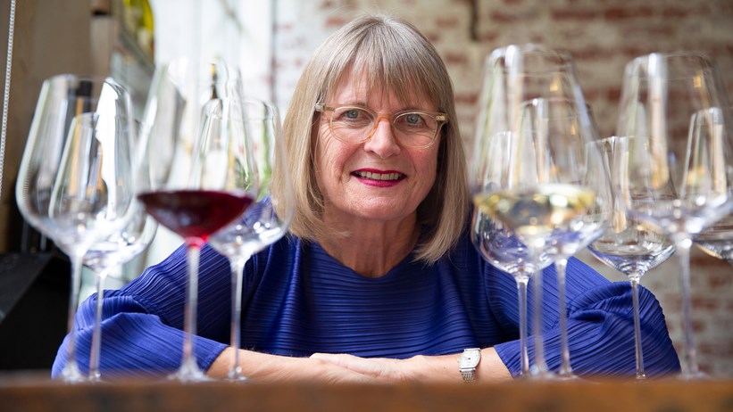 Image of Jancis Robinson she is sat a table with both hand on the table. Either side of her is a number of wine glasses one of which is filled with red wine.